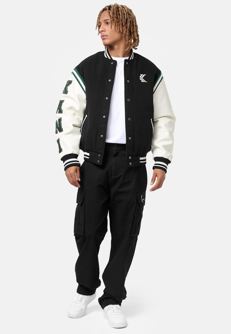 Jachetă varsity neagră și crem cu accente verzi, inscripții mari și manșete cu dungi, asortată cu pantaloni cargo negri și adidași albi.