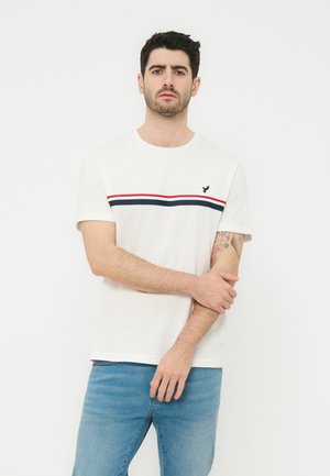 Sötét hajú férfi, fehér, piros és tengerészkék csíkos pólóban és kék farmerben, fehér háttér előtt áll, bal karját jobb kezével tartva.