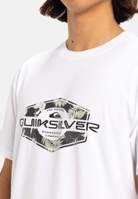 Camiseta blanca con un gráfico hexagonal que incluye texto negro y un patrón floral que rodea las palabras "Quiksilver Boardriding Company".