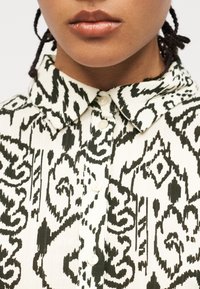 Κοντινό πλάνο ενός ατόμου που φοράει λευκό πουκάμισο με κουμπιά, με μαύρο αφηρημένο γεωμετρικό σχέδιο και μυτερό γιακά.