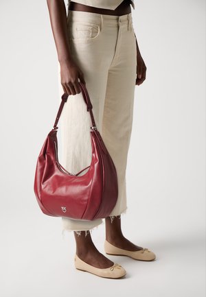 Woman holding a large red leather handbag, wearing off-white cropped jeans and beige ballet flats against a plain white background.