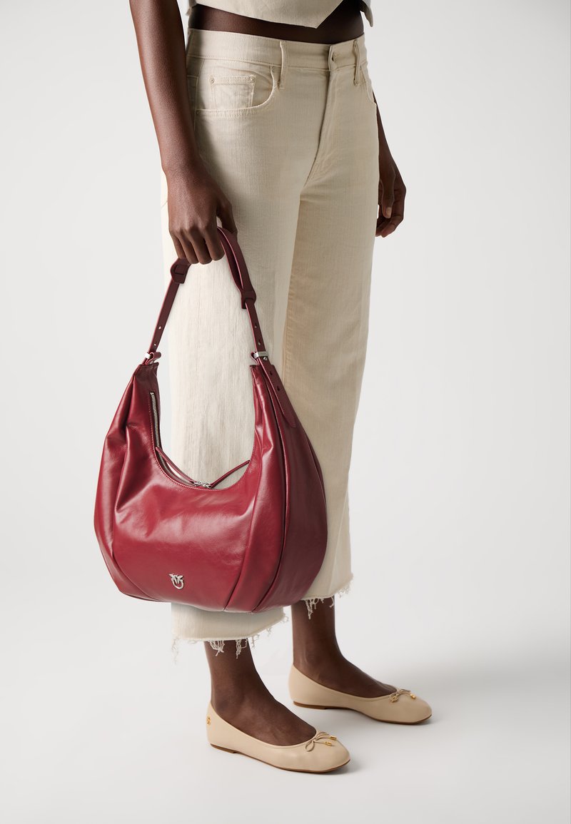 Woman holding a large red leather handbag, wearing off-white cropped jeans and beige ballet flats against a plain white background.