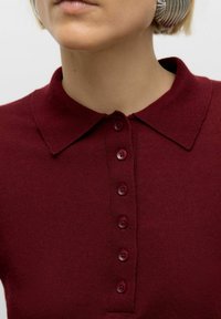Close-up of person wearing a burgundy buttoned collared shirt and large silver hoop earring against a plain background.