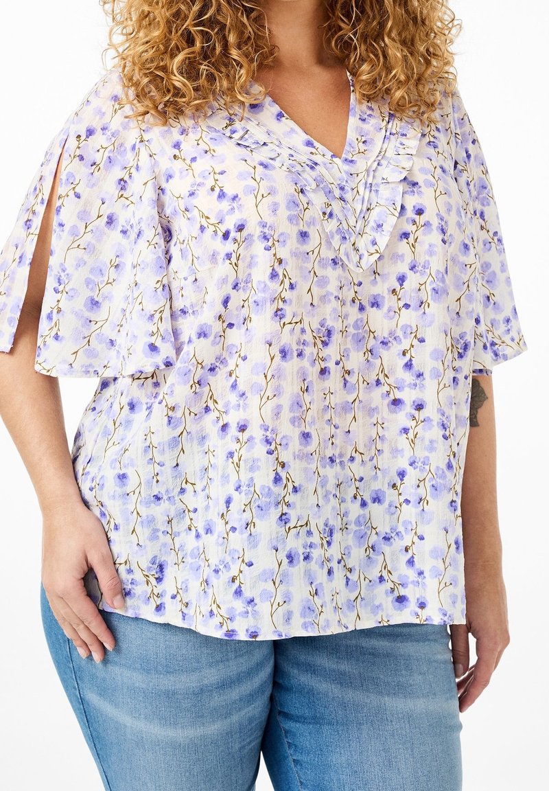 Blouse à motif floral avec un fond lavande clair, un col volanté et des manches courtes avec des découpes. Tissu doux et texturé.