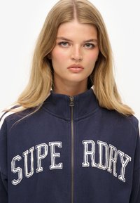Chaqueta de cremallera azul marino con el texto bordado en blanco "SUPERDRY", cuello alto y rayas blancas contrastantes en los hombros. Corte clásico.