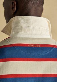 Mies, joka on pukeutunut raidoitettuun rugby-paitaan, jossa on nostettu vaalean beige kaulus. Paidassa on vaakasuoria punaisia, sinisiä ja vaalean beigejä raitoja, ja takana on ommeltu "JOULES".