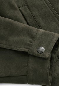 Olive green, suede jacket detail featuring a button closure, smooth texture, and stitched seams. Includes a side pocket for functionality.