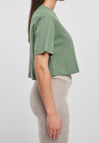 Zkrácená tričko s krátkým rukávem v tlumené zelené barvě z bavlny s kulatým výstřihem, zobrazené z boku, spárováno s světle béžovými kordurovými kalhotami.