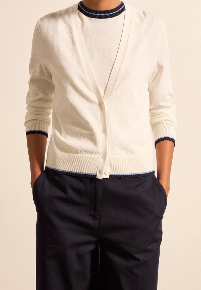 Personne portant un cardigan blanc boutonné par-dessus une chemise blanche assortie avec un bord bleu marine, associée à un pantalon bleu marine foncé, les mains dans les poches.