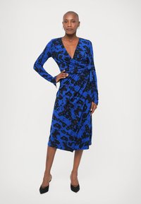 Žena stojí s rukou na boku, oblečená v modro-čiernych vzorovaných zavinovacích šatách a čiernych podpätkoch, na pozadí bielej steny.