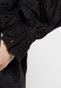 Sort stofærme med indviklet broderi af blomsterudskæringer og bølget blondekant på en persons arm mod en neutral baggrund.