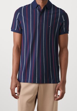 Man draagt een marineblauw poloshirt met verticale witte en rode strepen en beige broek, staand tegen een effen lichte achtergrond.