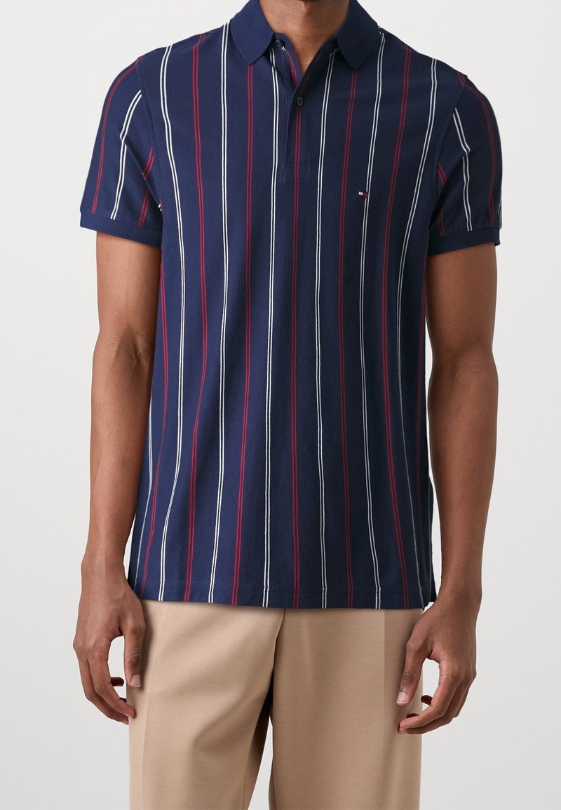 Homme portant un polo bleu marine avec des rayures verticales blanches et rouges, et un pantalon beige, debout devant un fond clair uni.