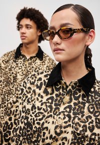 Barna és fekete kordbársony leopárdmintás ing fekete velúrból készült gallérral; a modellen teknősbéka mintás ovális napszemüveg van.