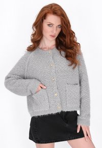 Žena s rezavými vlasy, oblečená v texturovaném šedém cardigan s knoflíky a černé džínové sukni, s jednou rukou v kapse cardigan, pózující proti bílému pozadí.