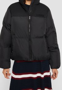 Chaqueta acolchada negra con cierre frontal de cremallera, diseño acolchado, cuello alto y parche de logo en el pecho. Textura lisa, corte corto.