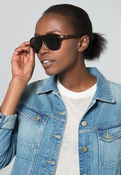 Occhiali da sole marrone tartaruga con lenti scure, montatura rettangolare e dettagli metallici, indossati da una persona con una giacca di jeans sopra un top in maglia bianca.