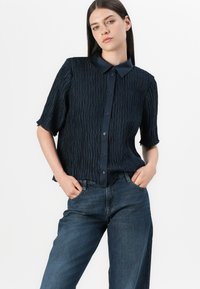Donkerblauwe shirt met korte mouwen, voorzien van een gestructureerd, geplooid ontwerp, knoopsluiting aan de voorkant en een klassieke kraag, gecombineerd met wijde denim jeans.