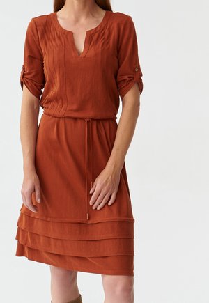 Kobieta w rdzawej sukience do kolan z podwiniętymi rękawami, ozdobiona przeszyciami z przodu oraz warstwowymi falbanami na spódnicy, stojąca na jednolitym tle.