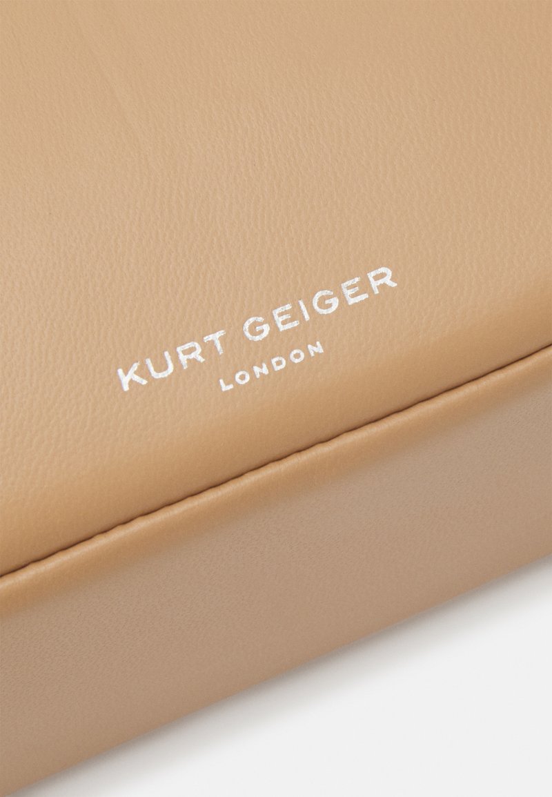 Beżowy skórzany dodatek o gładkiej fakturze, z wytłoczonym białym napisem "KURT GEIGER LONDON" na powierzchni.