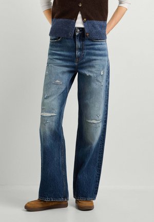 Person trägt hoch taillierte, weit geschnittene, abgenutzte blaue Jeans mit braunen Schuhen und einer geknöpften Weste über einem weißen Top, Hände in den Taschen.