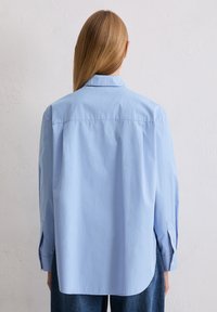 Jasnoniebieska koszula wykonana z gładkiego materiału, z klasycznym kołnierzykiem, długimi rękawami i lekko zakrzywionym dołem z tyłu.