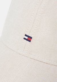 Beige cap with a textured fabric, featuring a small embroidered logo in navy, red, and white on the side.