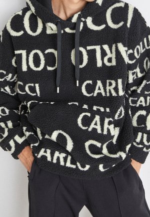 Zwart-witte fleece hoodie met patroon en grote herhaalde tekst, gedragen met zwarte broek, handen in zakken en zichtbare trekkoorden aan de capuchon.