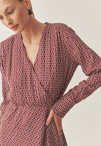 Piros mintás ruha V-nyakkivágással, hosszú ujjakkal és megkötős derékrésszel, fehér és kék mintákkal. Simaság érzetű anyag.