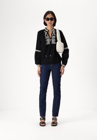 Μαύρη μπλούζα με μακριά μανίκια και λευκή floral κέντημα, συνδυασμένη με μπλε τζιν και μαύρες σαγιονάρες με χρυσά μεταλλικά στοιχεία; περιλαμβάνεται ώμου τσάντα.