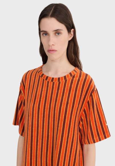 Mlada žena s dugom smeđom kosom koja nosi široku narančastu majicu s vertikalnim smeđim i žutim prugama, neutralnog izraza lica, siva pozadina.