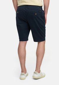Shorts bleu marine en tissu léger, dotés de deux poches arrière avec boutons et d'un ourlet droit au niveau du genou. Baskets blanches décontractées.