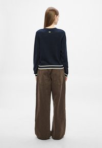 Ναυτικό πουλόβερ με ριμπ ύφανση και λευκές ρίγες στη βάση, συνδυασμένο με καφέ παντελόνια φαρδιάς γραμμής με πίσω τσέπες.