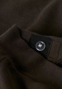 Tela texturizada de color marrón oscuro con un patrón acanalado. Presenta un botón claro en el cuello, sujeto a una lengüeta de tela negra.
