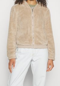 Frau trägt eine beige, flauschige Jacke mit Reißverschluss und hellblaue, hoch taillierte Jeans, steht vor einem schlichten hellen Hintergrund.