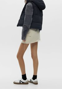 Svart pufferväst med huva över en grå långärmad tröja, kombinerad med en beige minikjol och vita sneakers med grå detaljer.