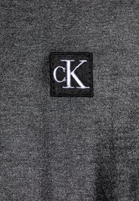 Šedá látka s texturovaným povrchem, obsahující obdélníkovou černou nášivku s bílým vyšitým logem "cK".