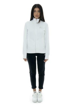 Une femme se tient devant un fond blanc, vêtue d’une veste blanche zippée, d’un pantalon noir et de baskets blanches.