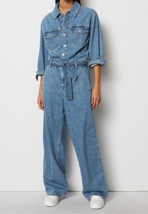 Frau trägt einen hellblauen Denim-Overall mit knöpfbarer Vorderseite, Brusttaschen, hochgekrempelten Ärmeln, gebundenem Gürtel und weißen Sneakers, steht vor einer Wand.