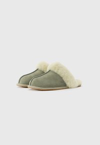 Zachte groene suède slippers met een crèmeachtige fluffy voering en tan rubberen zolen, naast elkaar geplaatst op een witte achtergrond.