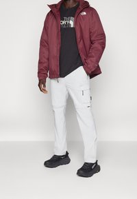 Burgundská bunda s kapucí přes černé tričko s logem, spárovaná s bílými cargo kalhotami a černými sportovními botami. Hladké textury, moderní střih.