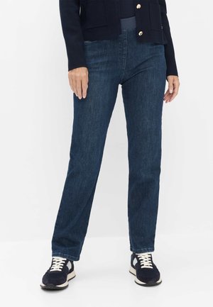 Person iført mørkeblå straight-leg jeans, sort jakke med guldknapper og sorte sneakers med hvide snørebånd, stående på et hvidt gulv.