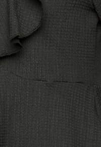 Ciemnozielona, teksturowana tkanina z pofałdowanym wzorem, z falbanami na krawędziach przy dekolcie, podkreślającymi jej warstwowy design.