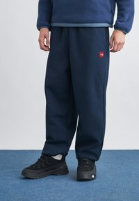 Άτομο που φοράει σκούρο μπλε φόρμα με κόκκινο λογότυπο, μαύρα αθλητικά παπούτσια, λευκές κάλτσες και μπλε φλις μπουφάν, στέκεται σε μπλε μοκέτα.