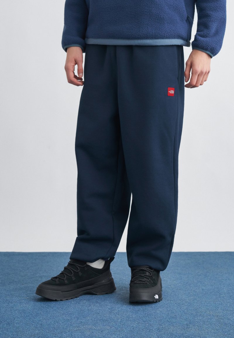 Άτομο που φοράει σκούρο μπλε φόρμα με κόκκινο λογότυπο, μαύρα αθλητικά παπούτσια, λευκές κάλτσες και μπλε φλις μπουφάν, στέκεται σε μπλε μοκέτα.