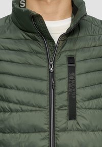 Grüne, fluffige Jacke mit horizontalem Steppmuster, hohem Kragen, vorderem Reißverschluss und einem schwarzen Textil-Detail mit Markenlogo auf der linken Seite.