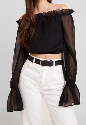 Frau trägt schwarzen durchsichtigen schulterfreien Crop-Top mit Rüschensäumen und weiße hoch taillierte Hose mit schwarzem Gürtel und goldener Schnalle.