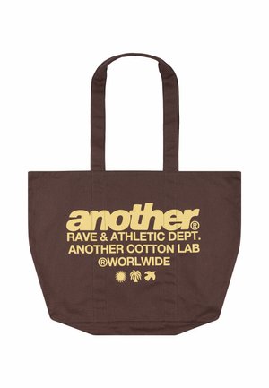 Brązowa torba na ramię z długimi uchwytami, żółty napis „another RAVE & ATHLETIC DEPT. ANOTHER COTTON LAB WORLDWIDE” i trzy małe ikony pod tekstem.