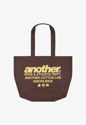 Brązowa torba na ramię z długimi uchwytami, żółty napis „another RAVE & ATHLETIC DEPT. ANOTHER COTTON LAB WORLDWIDE” i trzy małe ikony pod tekstem.