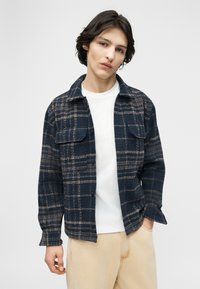 Νεαρό άτομο με μεσαίου μήκους σκουρόχρωμα μαλλιά φοράει ένα ναυτικό καρό σακάκι πάνω από λευκό πουκάμισο και μπεζ παντελόνι, στέκεται μπροστά σε ένα απλό φωτεινό φόντο.